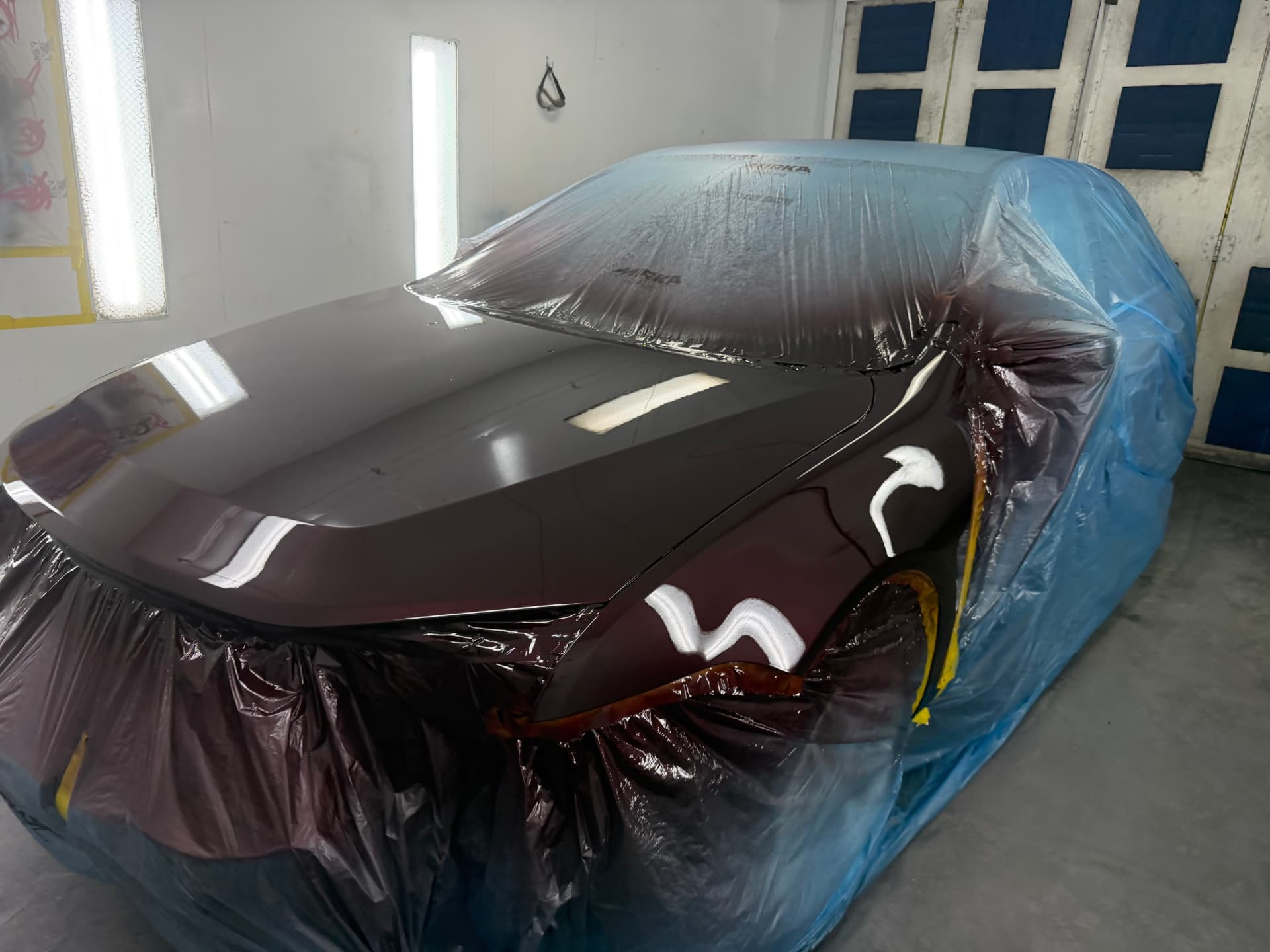 Sedan masked in plastic with a freshly sprayed deep maroon hood and fender in the paint booth